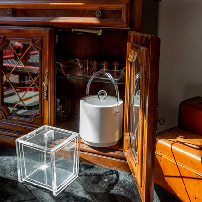 White ice bucket inside a wooden cabinet with a glass box on a dark surface.