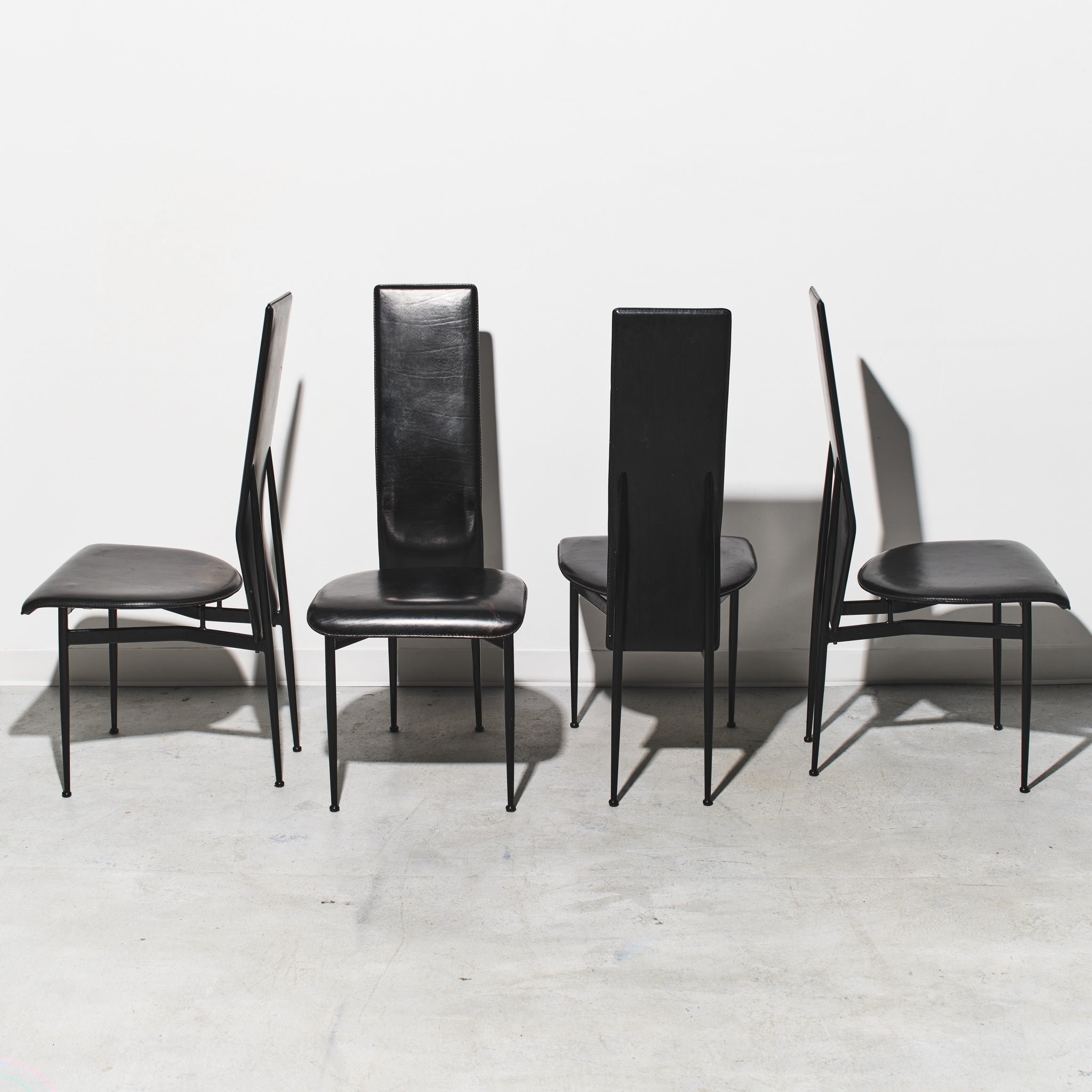 Four black chairs with thin legs on a light gray floor and white wall background