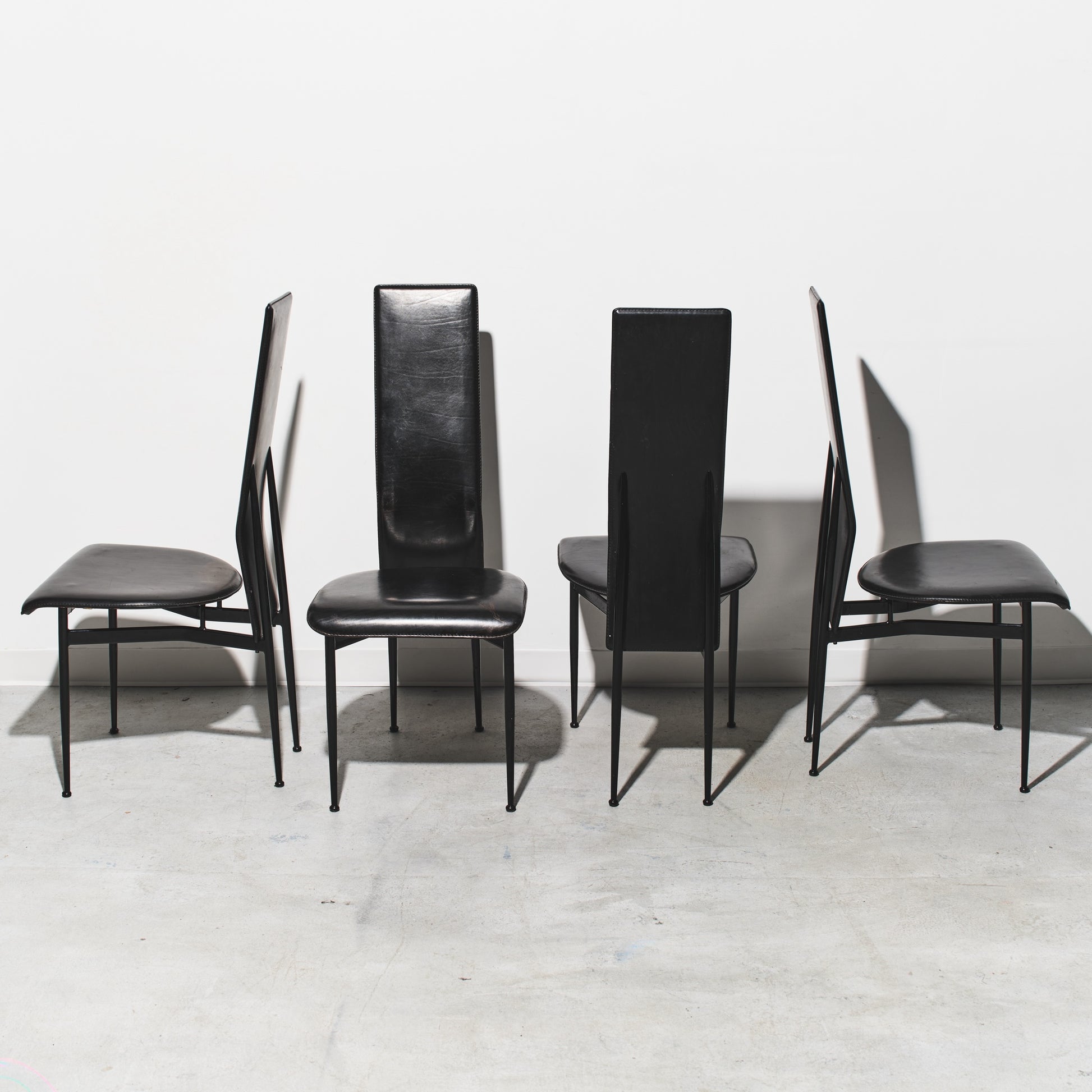 Four black chairs with thin legs on a light gray floor and white wall background