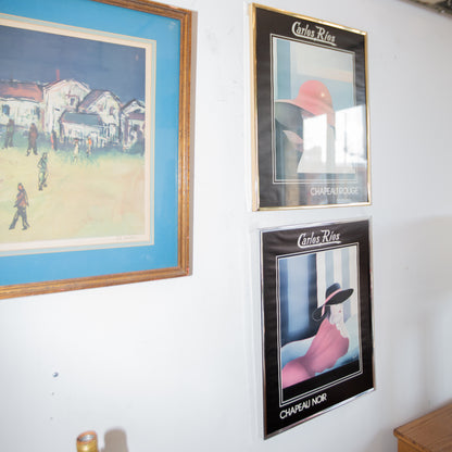 Three framed posters on a wall, featuring different scenes and a person with a hat.