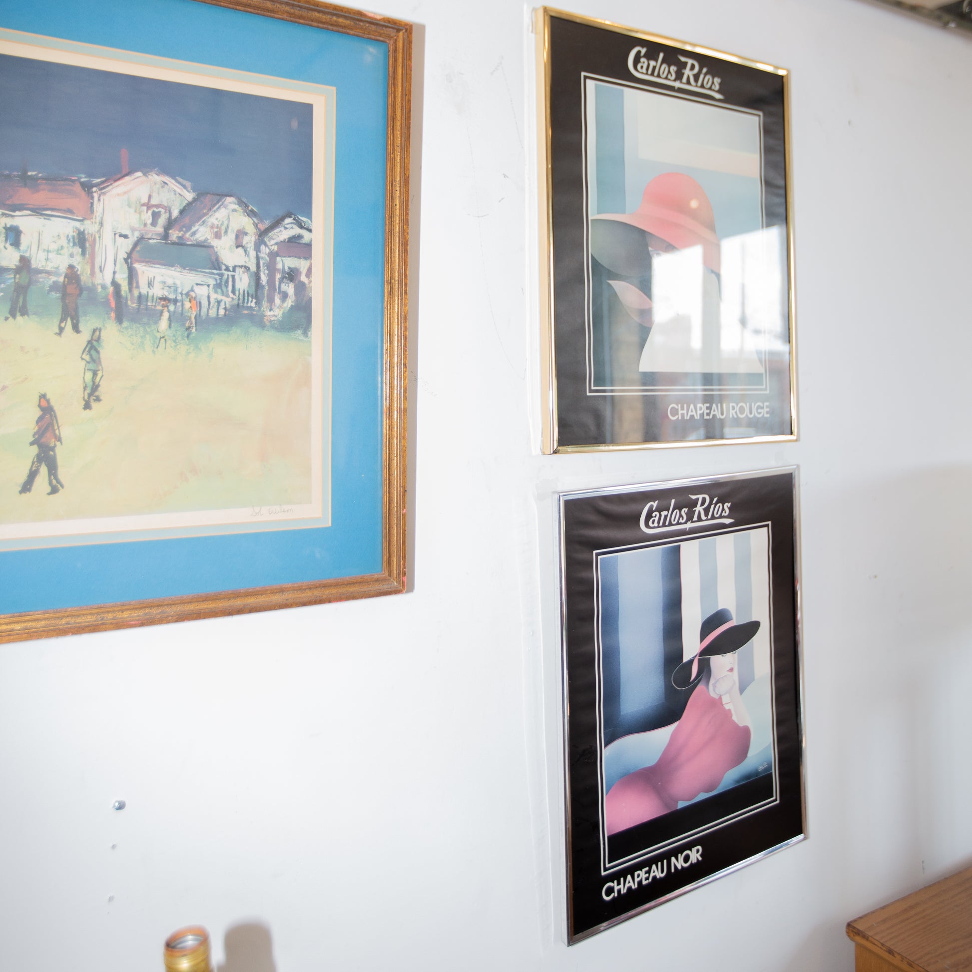 Three framed posters on a wall, featuring different scenes and a person with a hat.