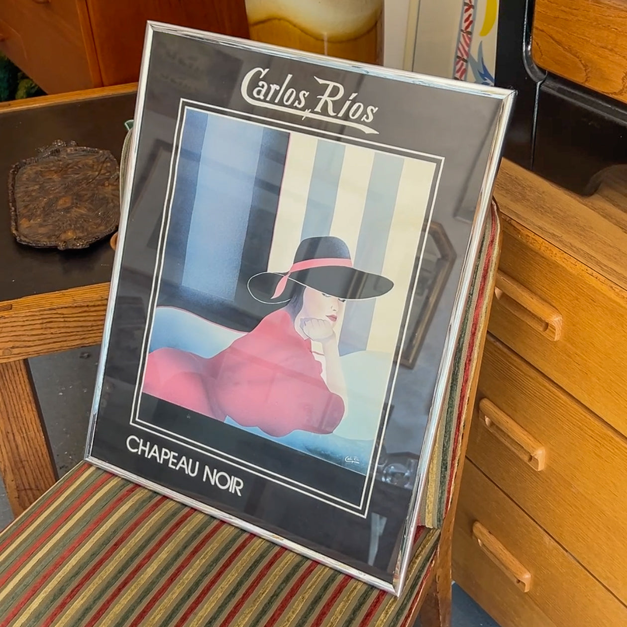 Framed artwork of a person wearing a black hat with 'Carlos Rios' branding on a striped surface.