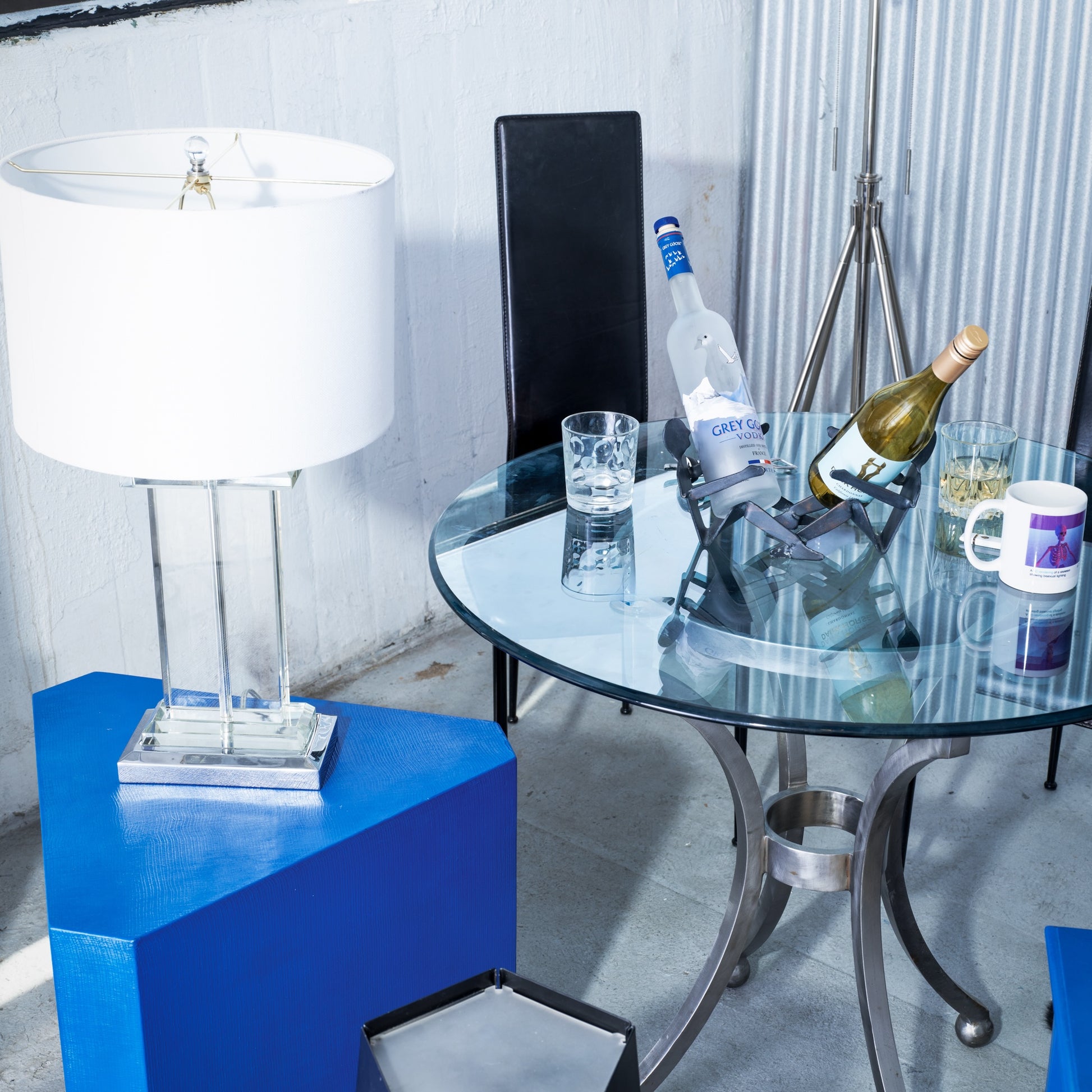 Glass table with drinks and a bottle on a blue side table against a white wall.