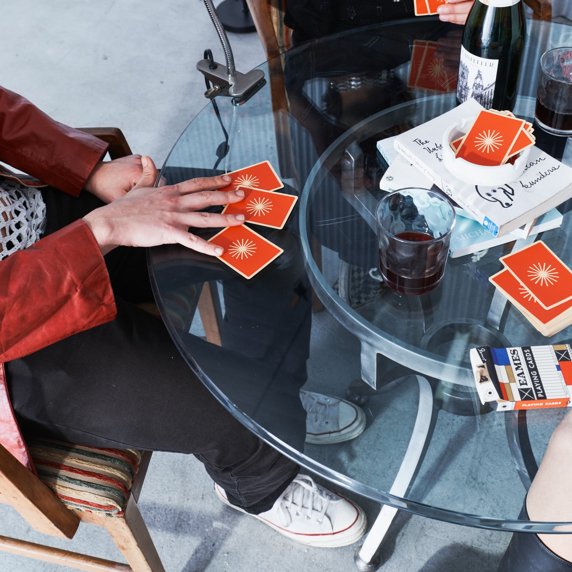 Two people sitting at a glass table with drinks and books, one person is reaching for a coaster.