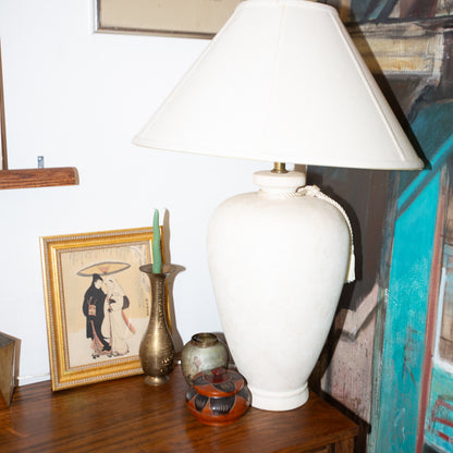 White lamp on a wooden surface with decorative items and a framed picture in the background.