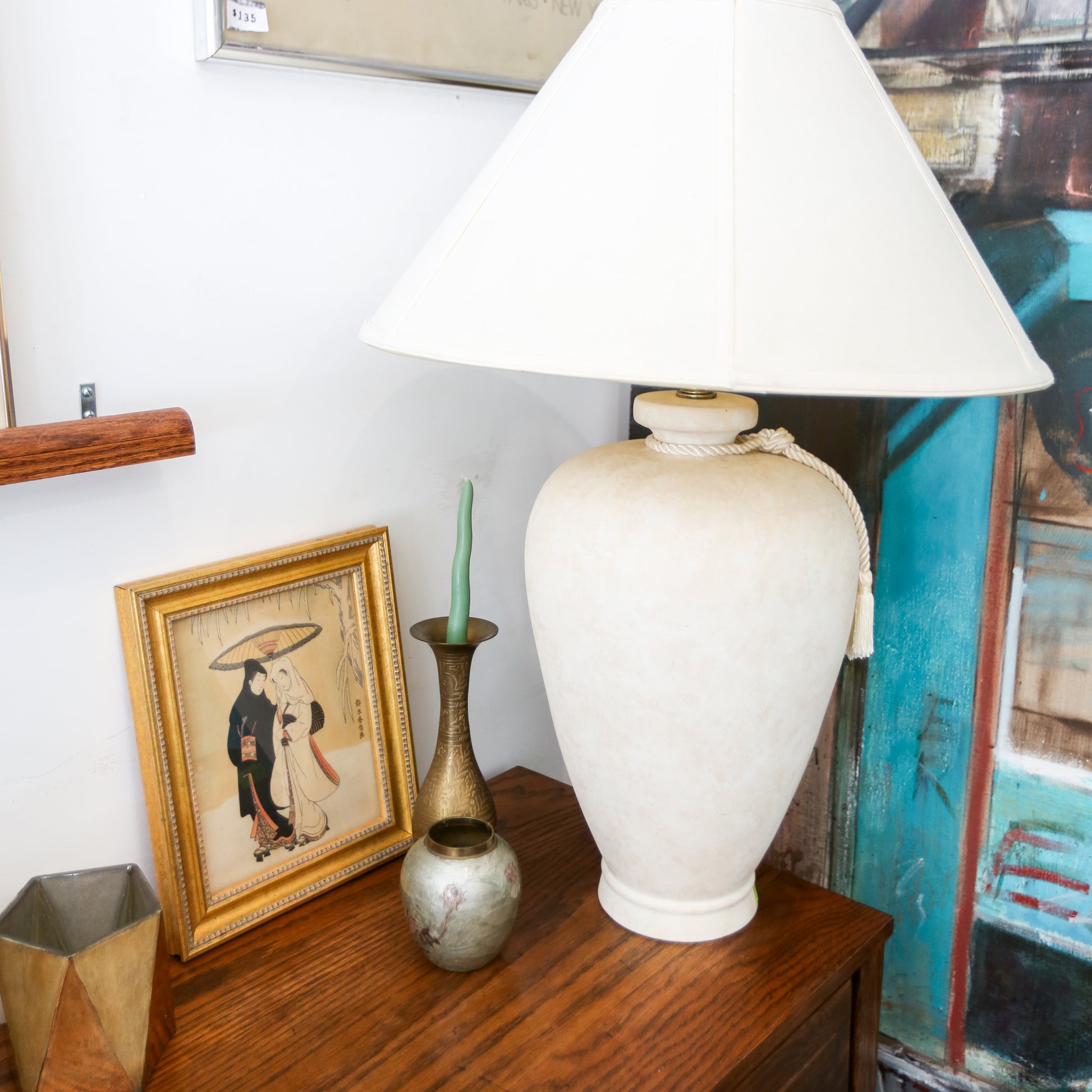 White lamp on a wooden surface with decorative items and a colorful painting in the background.