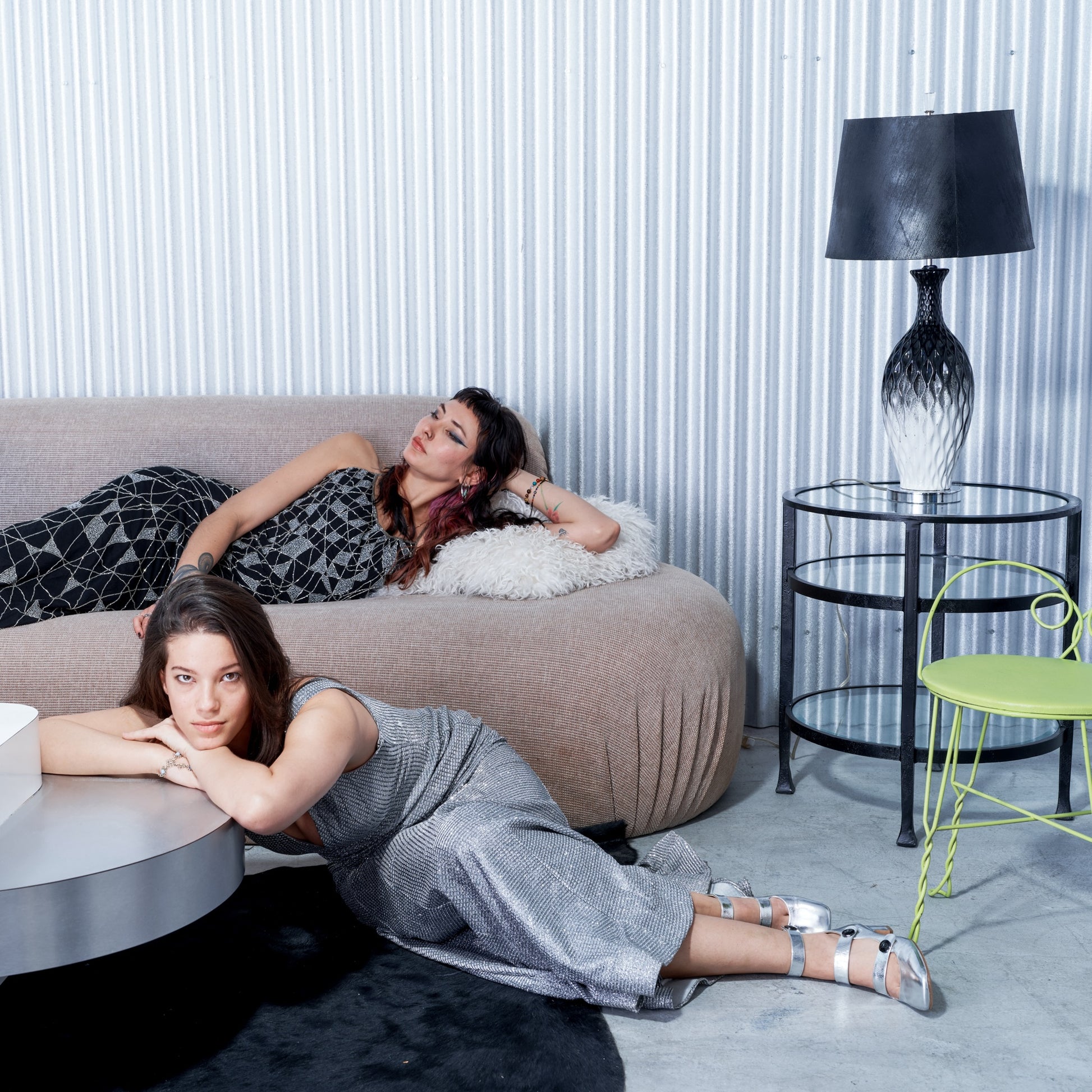 Two women lying on a couch in a modern living room with a lamp and chair.