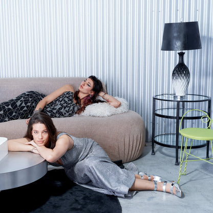 Two women lying on a couch in a modern living room with a lamp and chair.