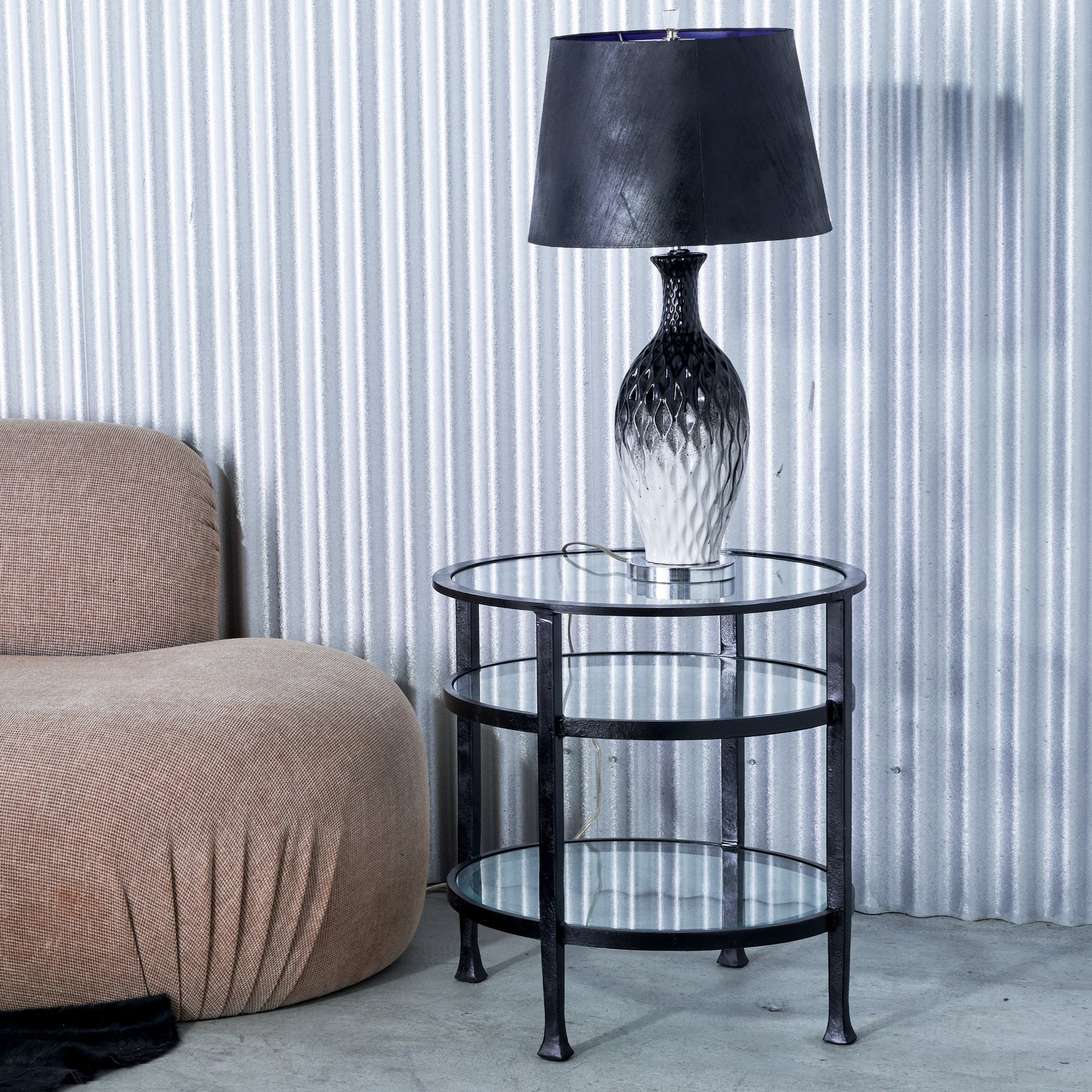 Round glass side table with a black lamp against a corrugated metal wall.