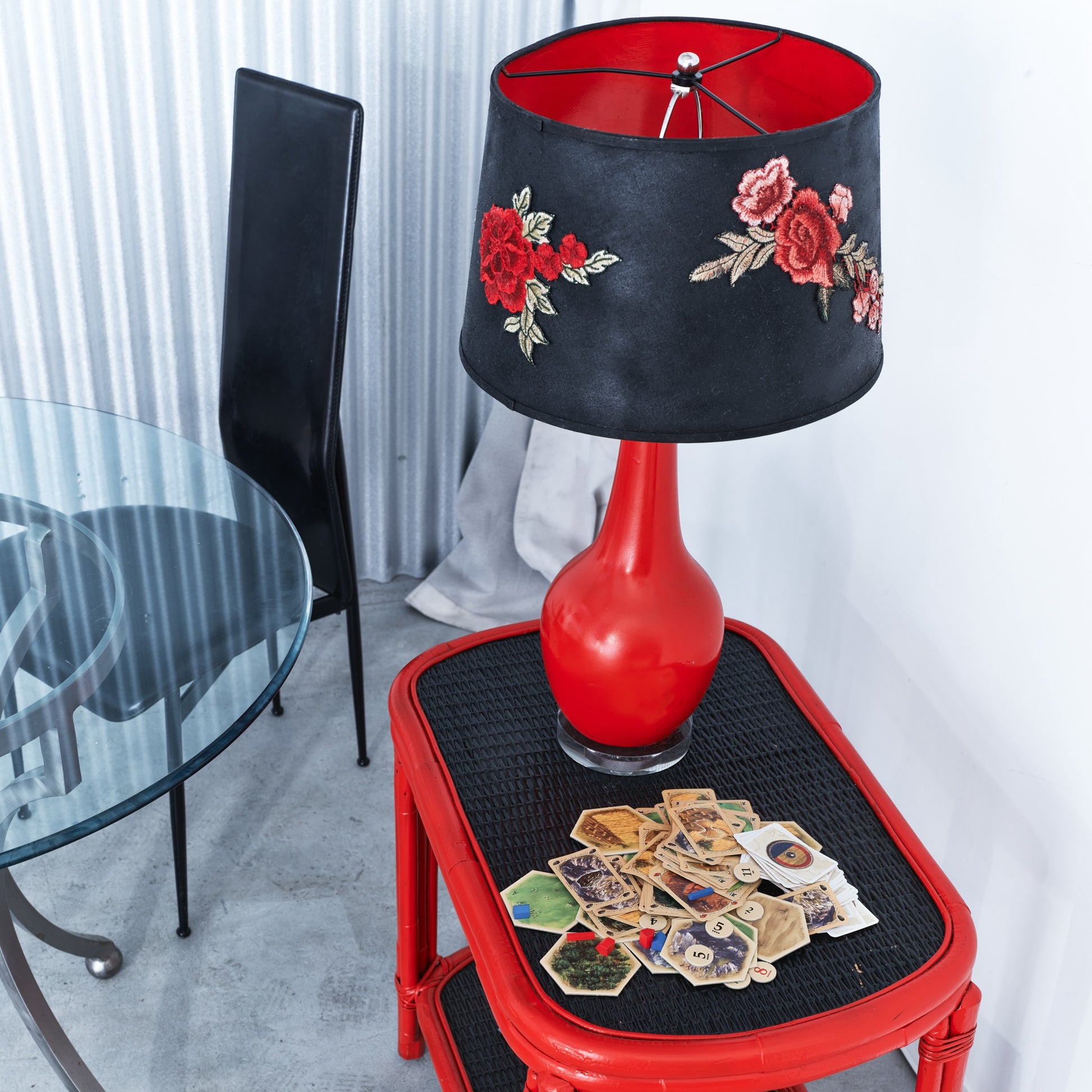 Red lamp with black floral shade on a red stool with playing cards, in a room setting.