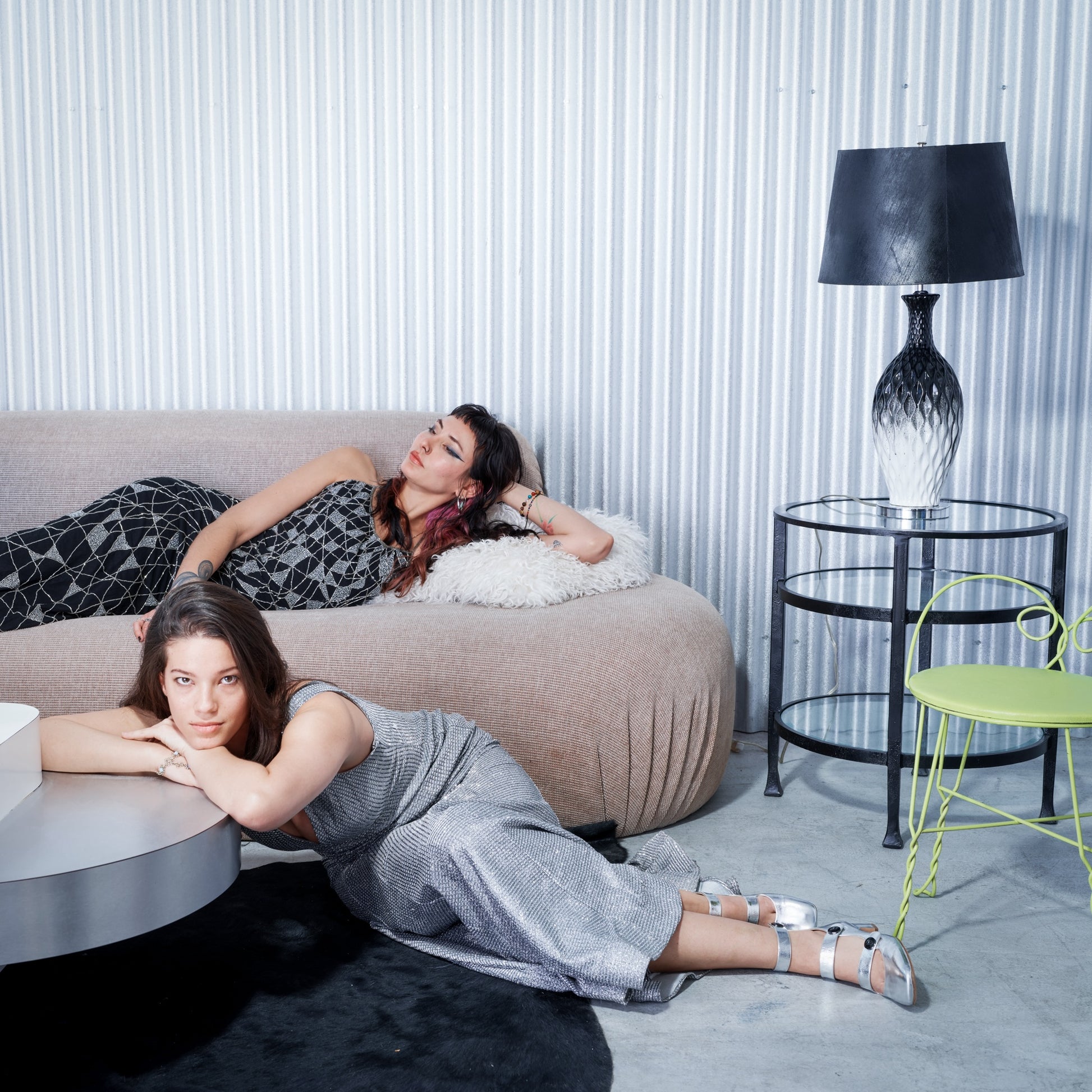 Two women lying on a beige sofa in a modern living room with a lamp and table.
