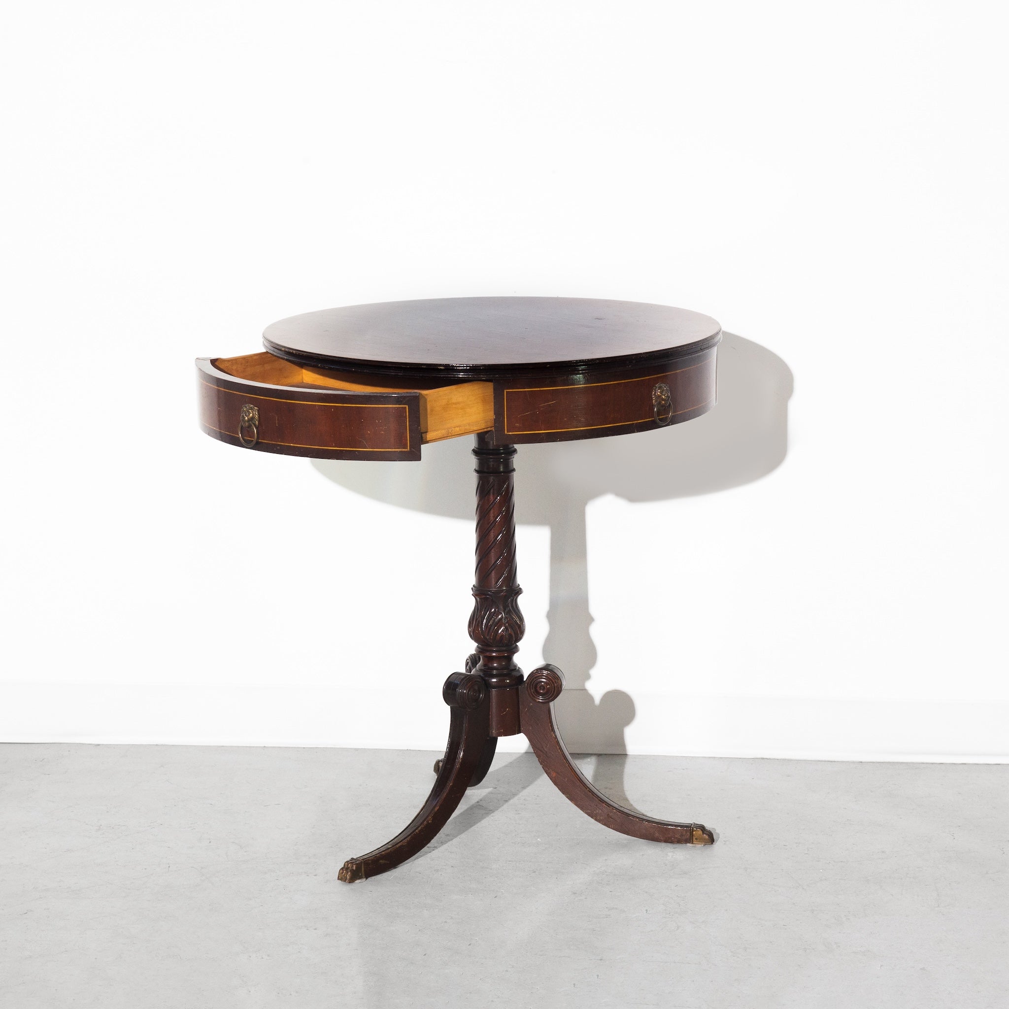 A round vintage wooden table with a central pedestal base and three feet features an open side drawer, set against a plain white wall and gray floor.